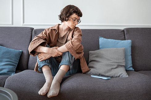 Young woman sitting on couch with knees held to chest while she looks at her cell phone and ignores the call