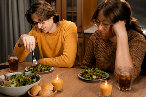 Man and woman sitting at dinner table both looking down awkwardly while eating salads
