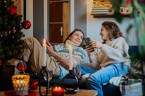 Two women sitting on couch looking at each other laughing and smiling surrounded by holiday decorations