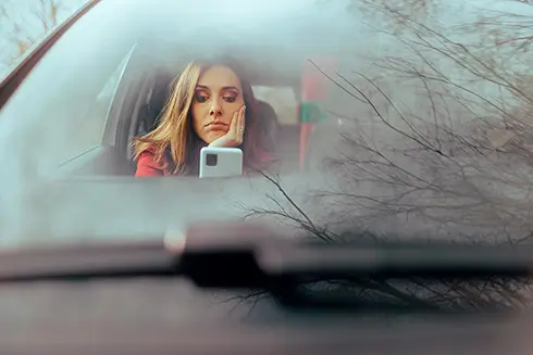 Woman sitting in car with windows frosted over looking at phone with disappointment