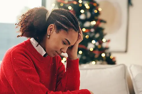 Woman in red holiday sweater in front of Christmas tree looking down with her head in her hand