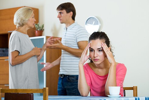 Woman sitting at table with hands on temples looking frustrated as her husband is talking to his overbearing mother in background